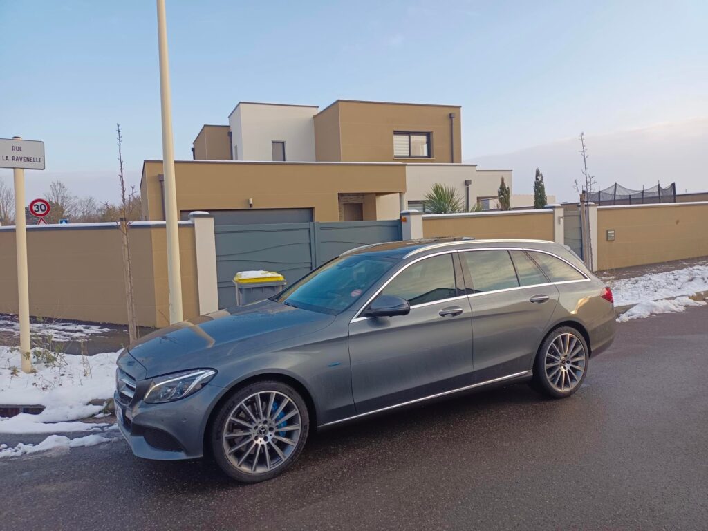 Break Mercedes gris stationné devant une maison avec de la neige au bord de la route.