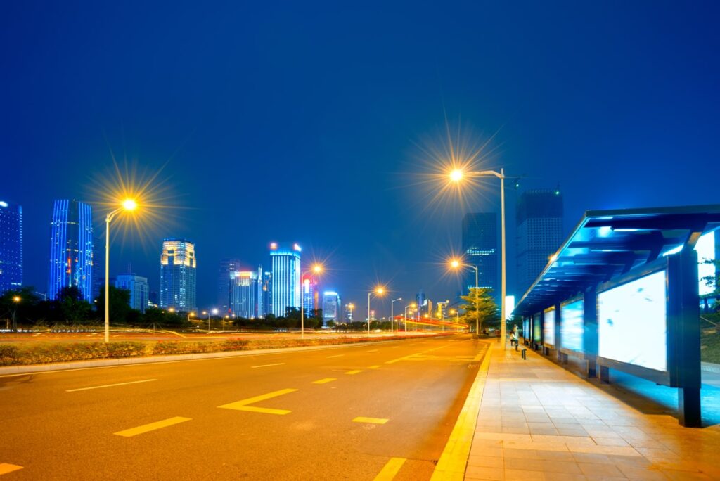 Route éclairée par des lampadaires avec un abribus et des immeubles illuminés de nuit.