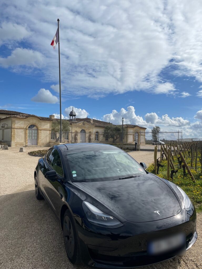 Tesla noire stationnée devant un domaine viticole et des rangs de vigne.