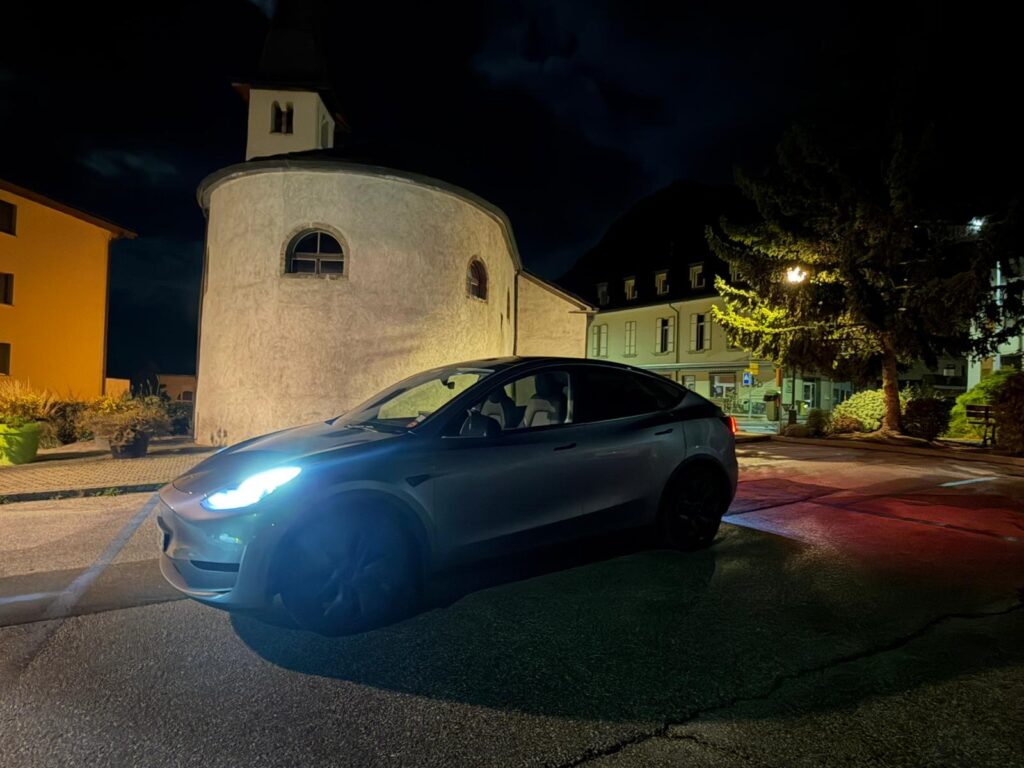 Voiture grise avec les phares allumés devant un bâtiment arrondi de nuit.