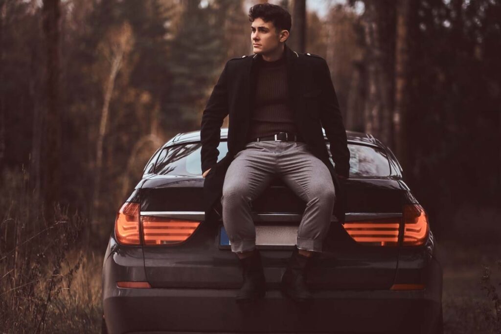 Homme assis sur le coffre d’une voiture noire dans un paysage boisé.
