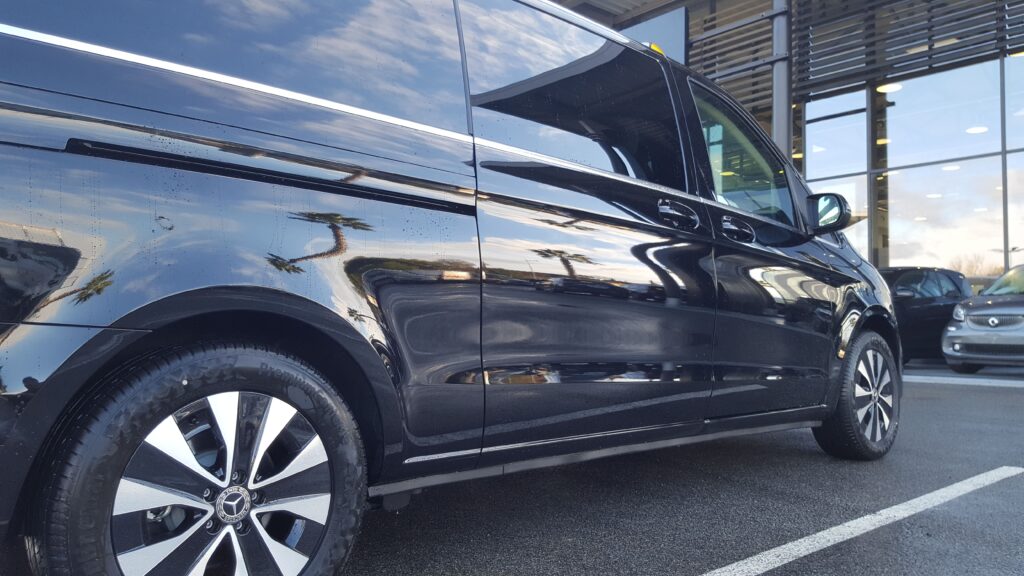 Vue latérale d’un van Mercedes noir stationné devant un bâtiment vitré.